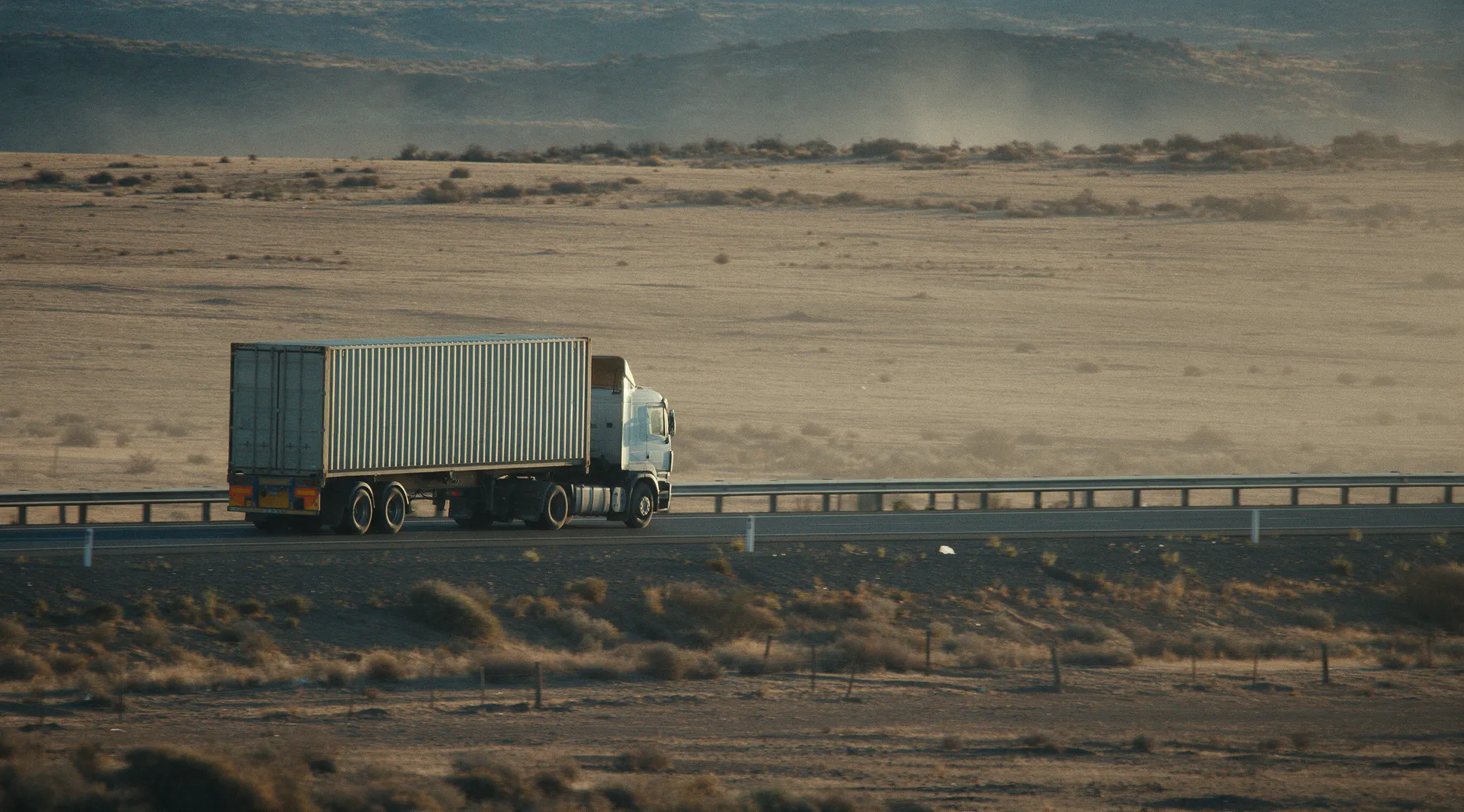 Camion porte-conteneur sur une autoroute marocaine traversant un plateau ocre, lumière rasante.