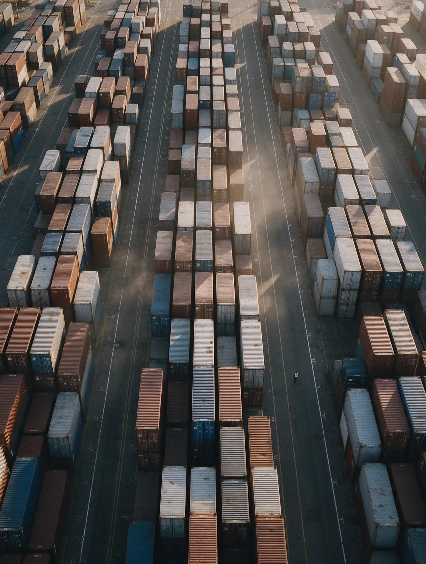 Rangées de conteneurs empilés vues du dessus dans un terminal portuaire, un ouvrier en gilet minuscule pour l'échelle.